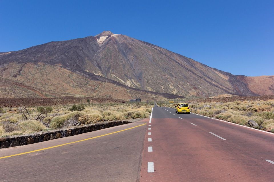 Una piccola auto gialla percorre una strada attraverso un paesaggio arido verso una grande montagna sotto un cielo azzurro chiaro.