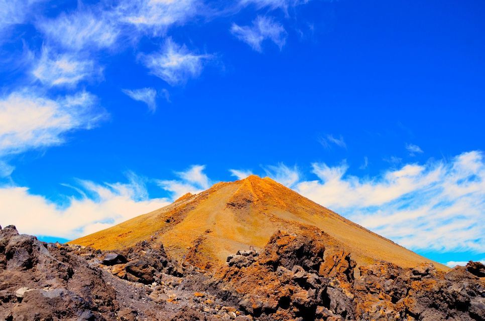 La cima arancione di una montagna vulcanica si erge sopra rocce laviche aspre sotto un cielo azzurro intenso con nuvole sottili.