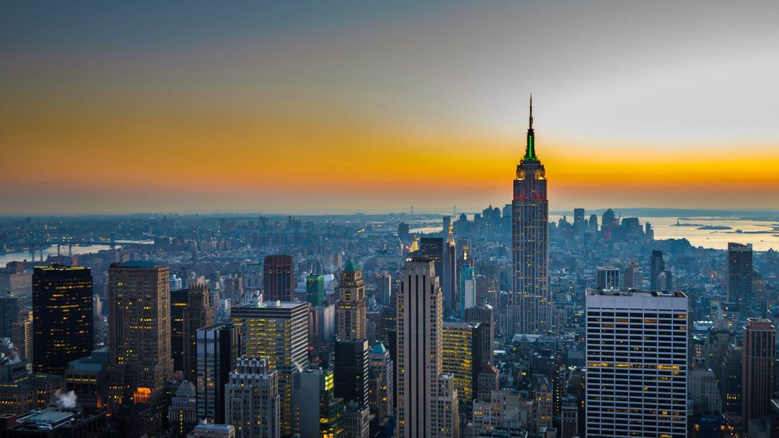 Un ampio skyline cittadino con numerosi grattacieli illuminati, inclusa una torre imponente, sullo sfondo di un vibrante tramonto.