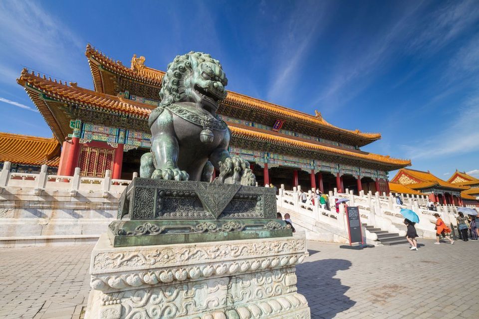 Une statue de lion gardien en bronze sur un piédestal dans une cour, avec un palais traditionnel au toit jaune en arrière-plan sous un ciel bleu.