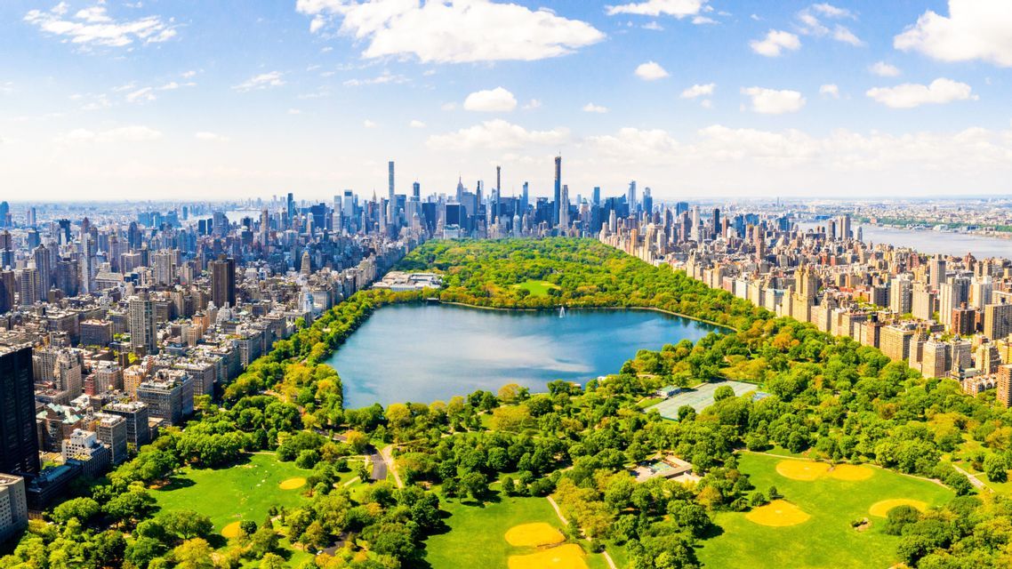 An aerial view of a large city park with a central lake, surrounded by skyscrapers and buildings on a sunny day.
