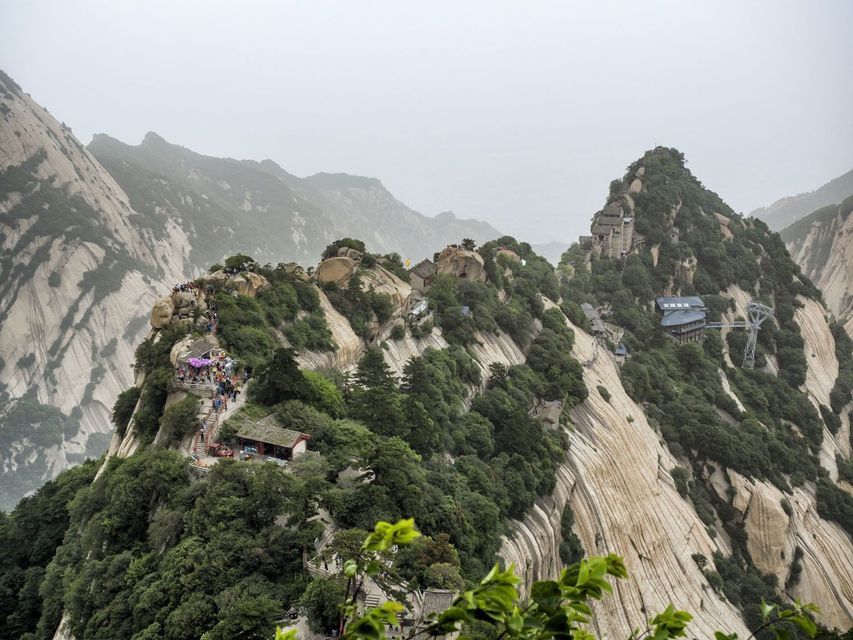 Una vista de una escarpada cordillera con picos rocosos, donde multitudes de personas caminan por un sendero entre templos y árboles verdes.