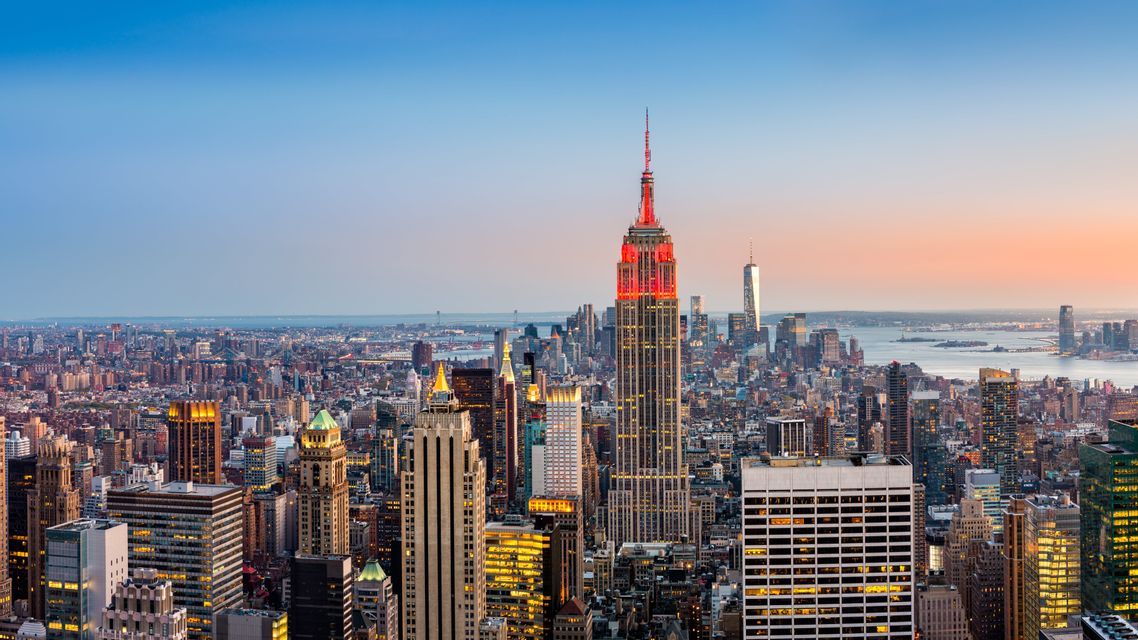 Eine Luftaufnahme der New Yorker Skyline in der Dämmerung, mit dem rot beleuchteten Empire State Building vor einem farbverlaufenden Himmel.