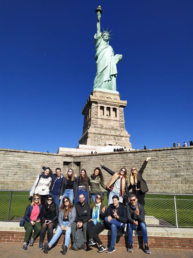 Eine WeRoad Gruppenreise posiert an einem sonnigen Tag für ein Foto vor einer großen grünen Statue einer Frau, die eine Fackel hält.