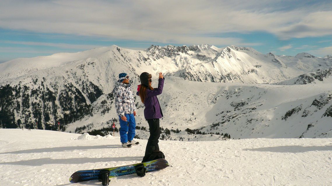 Zwei Snowboarder stehen auf einem verschneiten Berggipfel, einer fotografiert die weite, schneebedeckte Bergkette. Zwei Snowboarder stehen auf einem verschneiten Berggipfel, einer fotografiert die weite, schneebedeckte Bergkette.