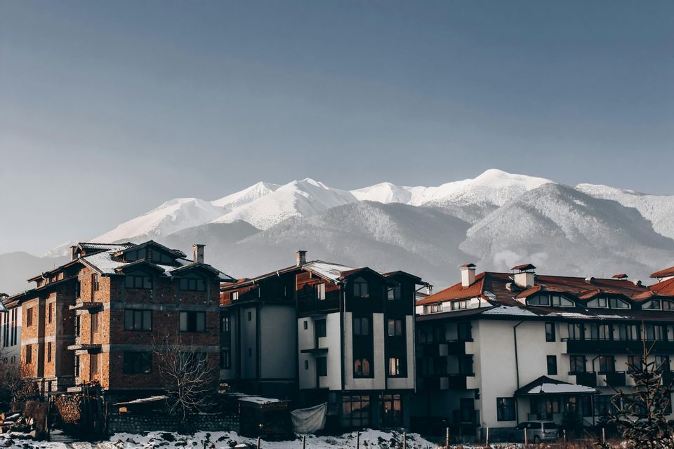 Edifici in stile alpino con tetti innevati si ergono di fronte a una vasta catena montuosa innevata sotto un cielo limpido.