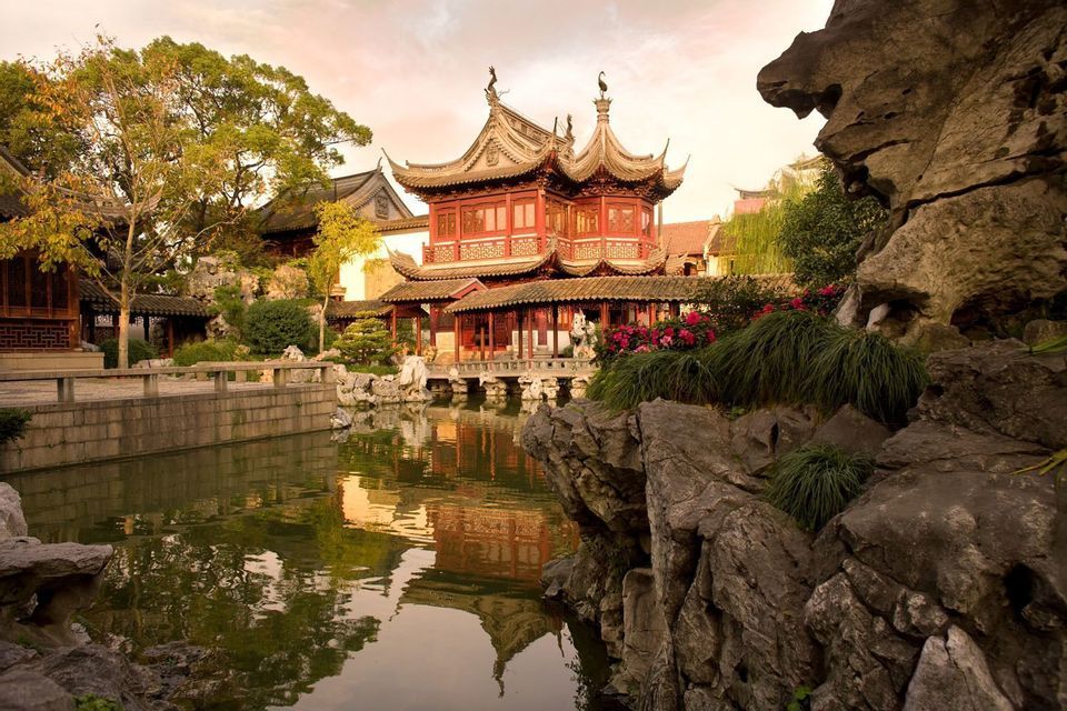 A traditional red Chinese pavilion with tiered roofs reflects in a calm pond, surrounded by lush gardens and rocks.