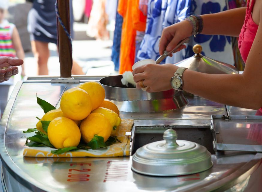 Un venditore ambulante dosa il sorbetto al limone da un carretto di metallo in una coppetta, accanto a una pila di limoni freschi sul bancone