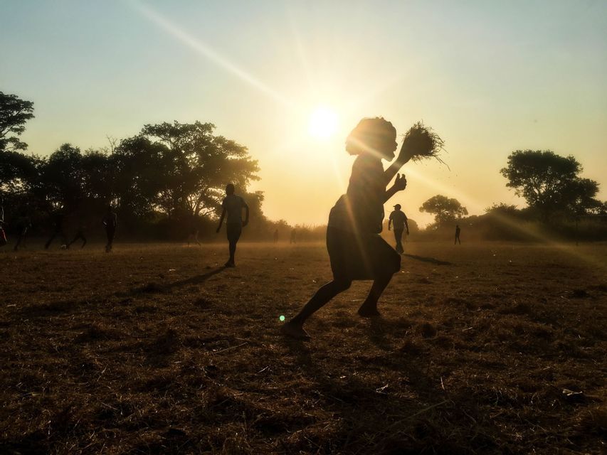 Una silhouette corre per un campo con una palla di paglia fatta a mano, mentre altri giocano sullo sfondo al tramonto.