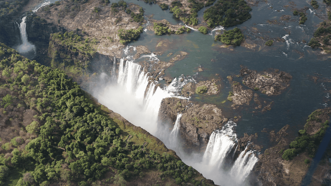 Veduta aerea di un vasto sistema di cascate, con l'acqua che precipita da una lunga scogliera in un profondo canyon fiancheggiato da fitta foresta verde.