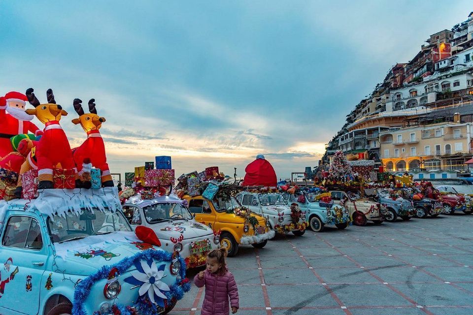 Una fila di Fiat 500 d'epoca adornate con elaborate decorazioni natalizie sono parcheggiate in una piazza al crepuscolo.