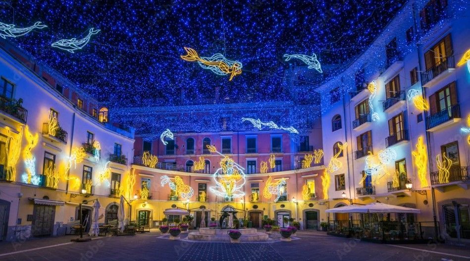 Una piazza cittadina di notte, illuminata da elaborate installazioni luminose di sirene, delfini e pesci sotto una tettoia di luce blu.