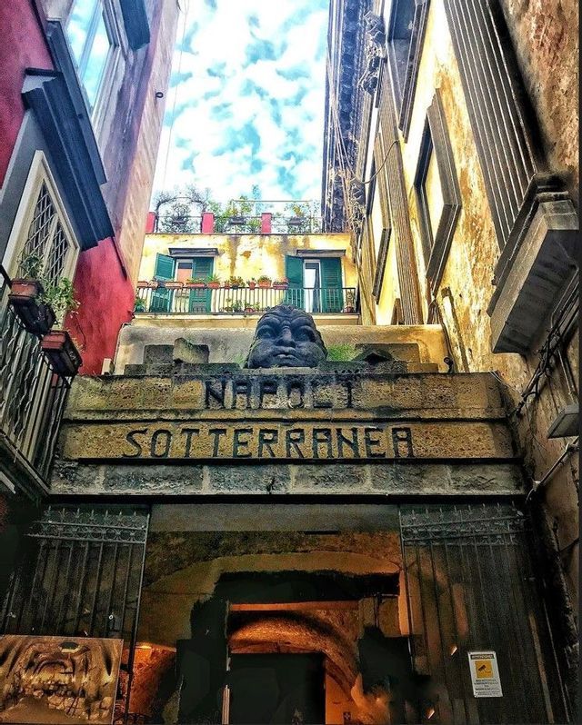 Vista en contrapicado de la entrada de piedra a Napoli Sotterranea, con un rostro tallado sobre el arco en un callejón estrecho.