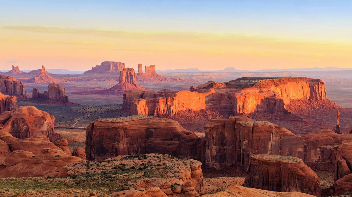 Un vasto paesaggio desertico con grandi formazioni rocciose rosse e mesa illuminate dalla calda luce di un sole nascente o al tramonto.