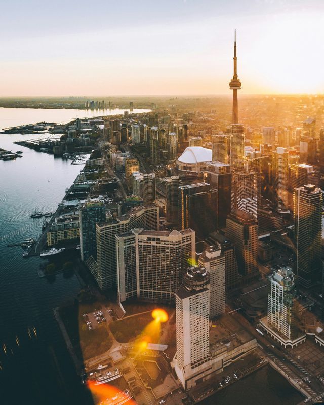 Una vista aerea di un fitto skyline cittadino con numerosi grattacieli e un'alta torre accanto a un grande specchio d'acqua al tramonto.