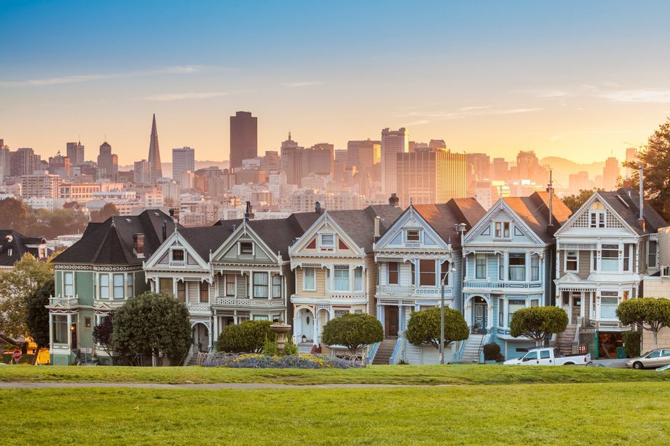A row of colorful Victorian houses with a modern city skyline in the background during sunset, seen from a grassy park.