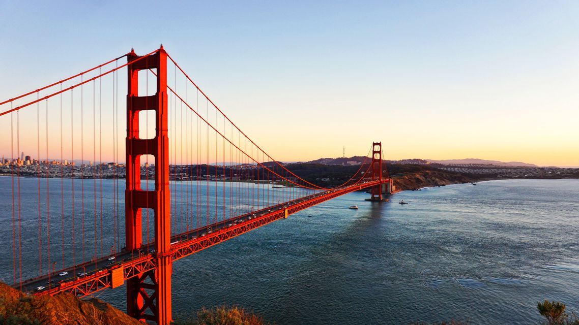 A large red suspension bridge spanning a bay at sunset, with cars crossing it and a city skyline in the background.