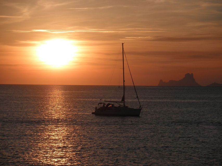 Un velero en silueta en el océano al atardecer, con el sol reflejándose en el agua y una isla a lo lejos.