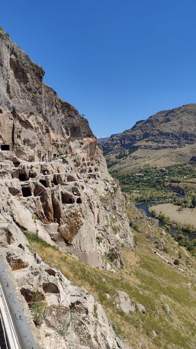 Abitazioni e grotte scavate in una grande scogliera si affacciano su una valle lussureggiante con un fiume sotto un limpido cielo azzurro.