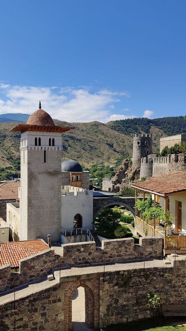 Una storica torre in pietra con tetto a cupola si erge all'interno di una città fortificata, con montagne verdi visibili sullo sfondo.