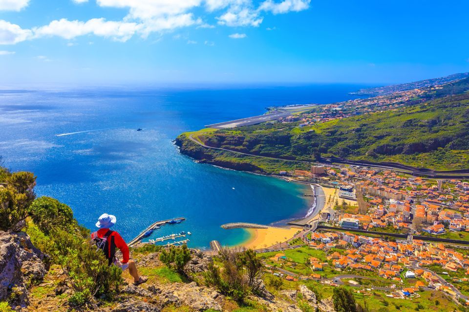 A person sits on a cliffside overlooking a coastal town with a sandy beach, a small harbor, and a vast blue sea.
