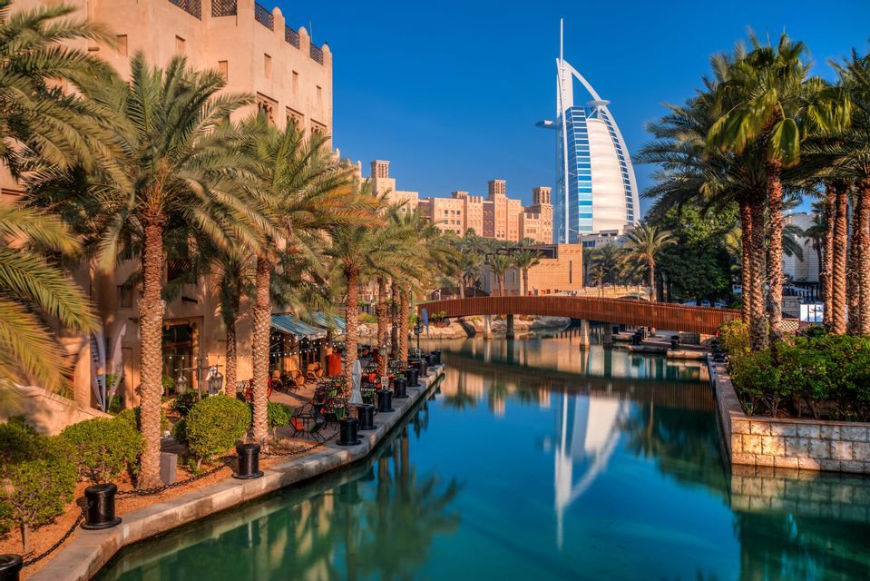 Un moderno grattacielo e edifici tradizionali riflessi in un canale fiancheggiato da palme in una giornata di sole.