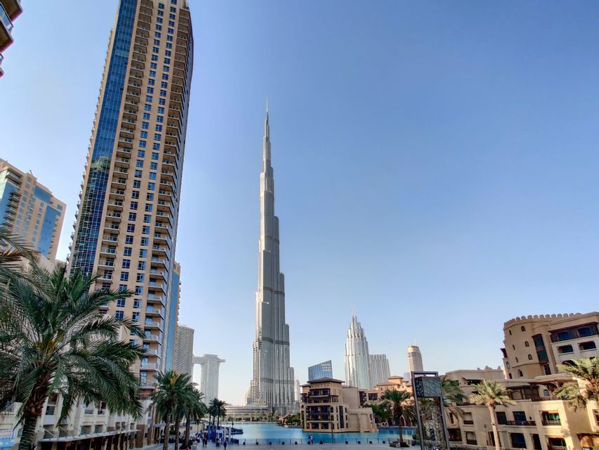 A tall, pointed skyscraper rises above a modern city with a large pool and palm trees in the foreground under a clear sky.