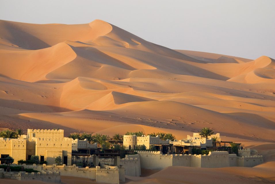 Un resort fortificato con palme è incastonato tra vaste e sinuose dune di sabbia arancione all'alba.