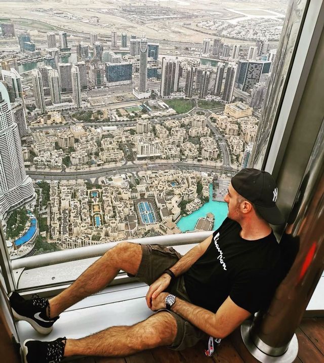 Un uomo con un berretto siede a terra vicino a una finestra, guardando un'ampia città da un punto di vista elevato.