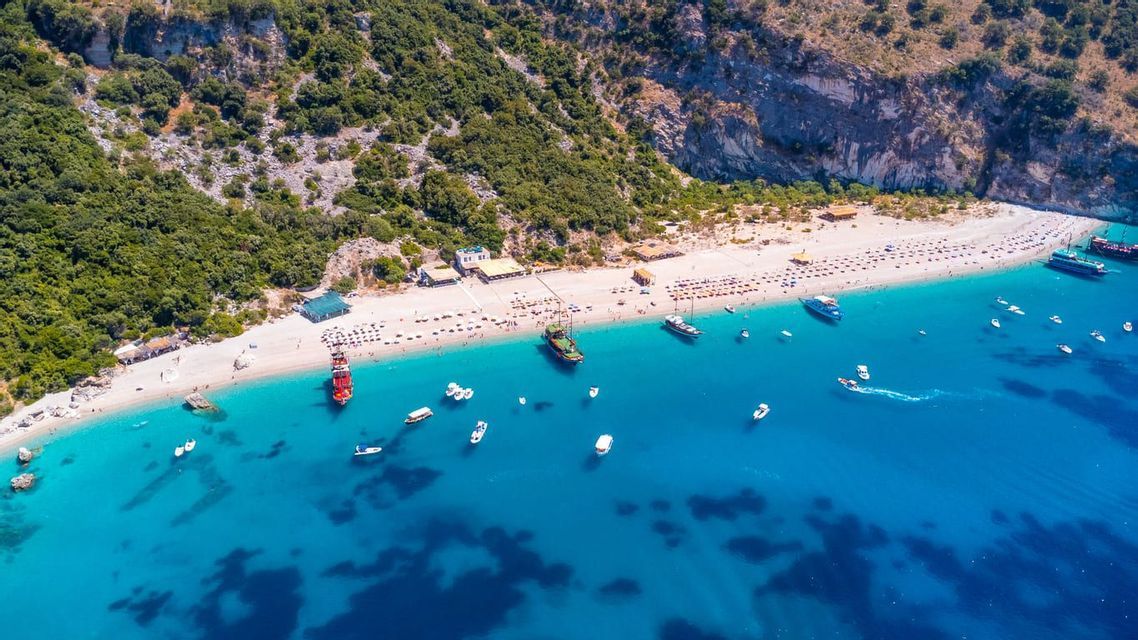 Una vista aerea di una spiaggia sabbiosa affollata con barche in acqua turchese, incastonata alla base di una collina rocciosa e boscosa.
