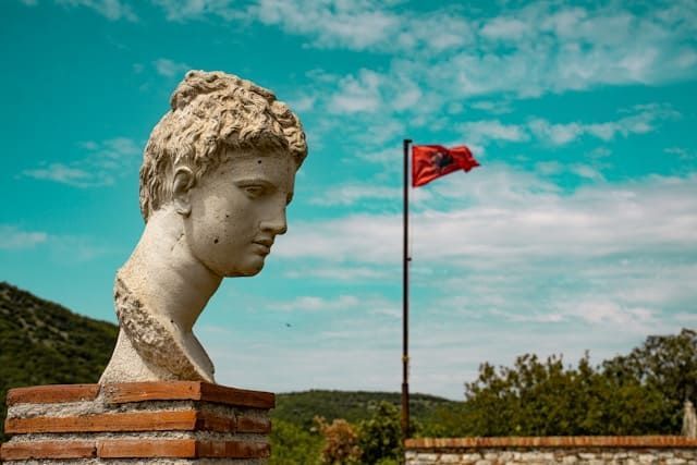 Un busto classico in pietra su un piedistallo di mattoni, con la bandiera albanese che sventola sullo sfondo contro un cielo turchese luminoso.