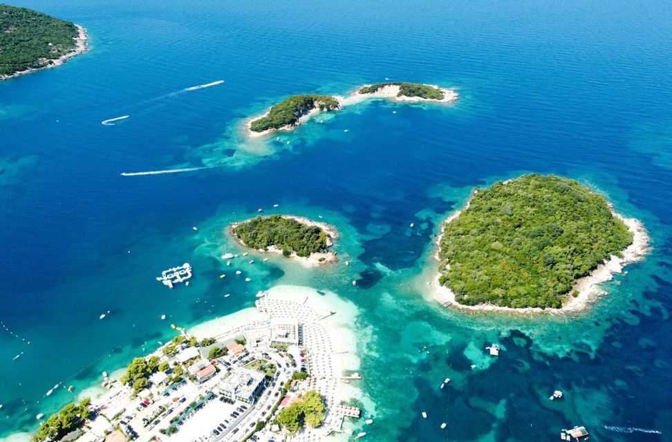 Una vista aerea di una costa con piccole isole verdi, una spiaggia affollata con ombrelloni e barche sul mare turchese.