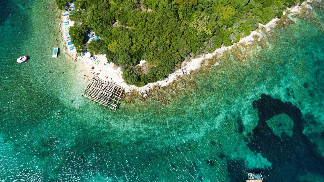 An aerial top-down view of a coastline with a small beach, people swimming in turquoise water, and a lush green forest.