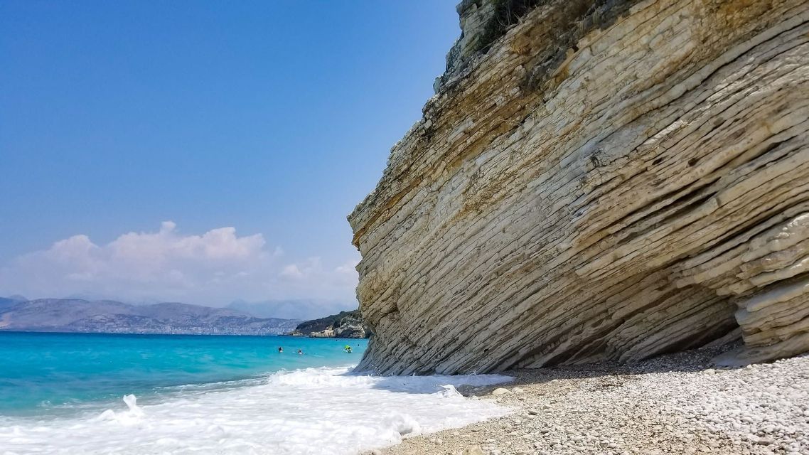 A large, layered rock cliff on a pebble beach with turquoise water and gentle waves under a clear blue sky.