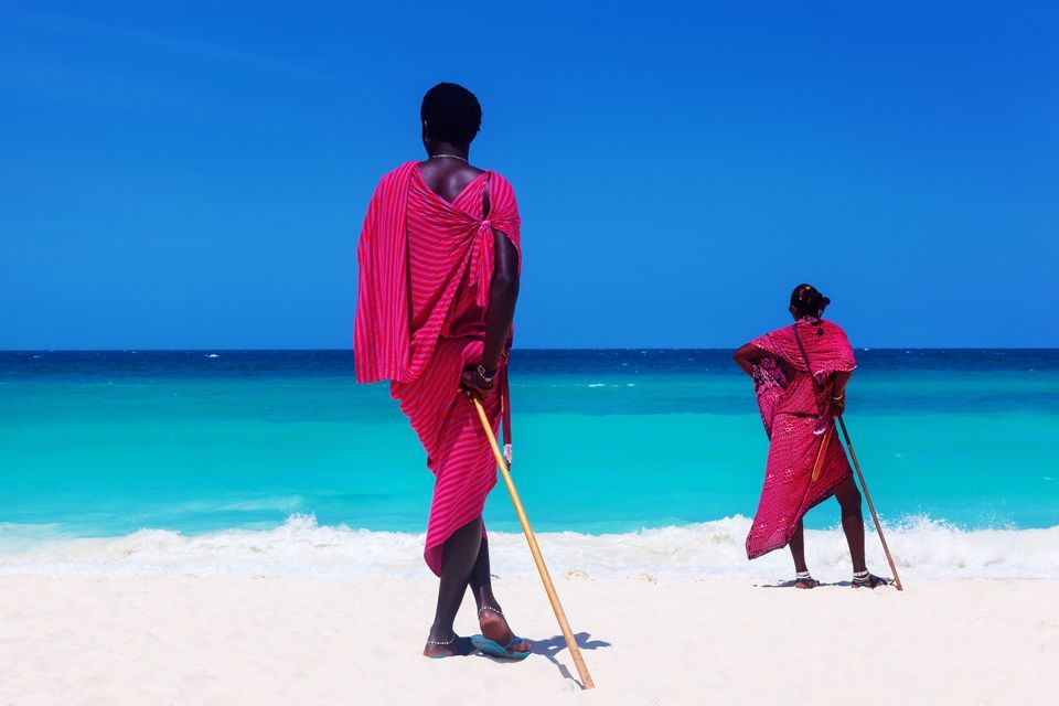 Due persone in abiti tradizionali rosa stanno in piedi con bastoni su una spiaggia di sabbia bianca, guardando l'oceano turchese.