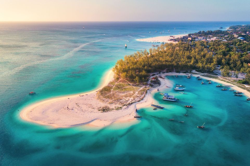 Vista aérea de una península de arena blanca que se extiende en agua oceánica turquesa, con muchos pequeños barcos anclados en una bahía al atardecer.