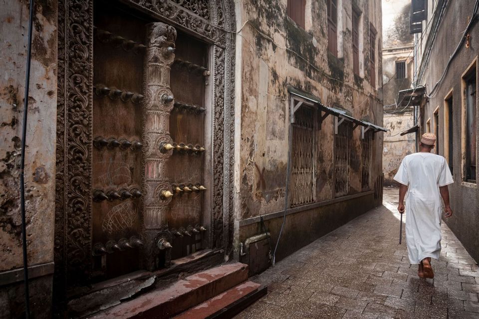 Un hombre con vestimenta tradicional blanca camina por un callejón estrecho y mojado junto a una gran puerta de madera ornamentada.