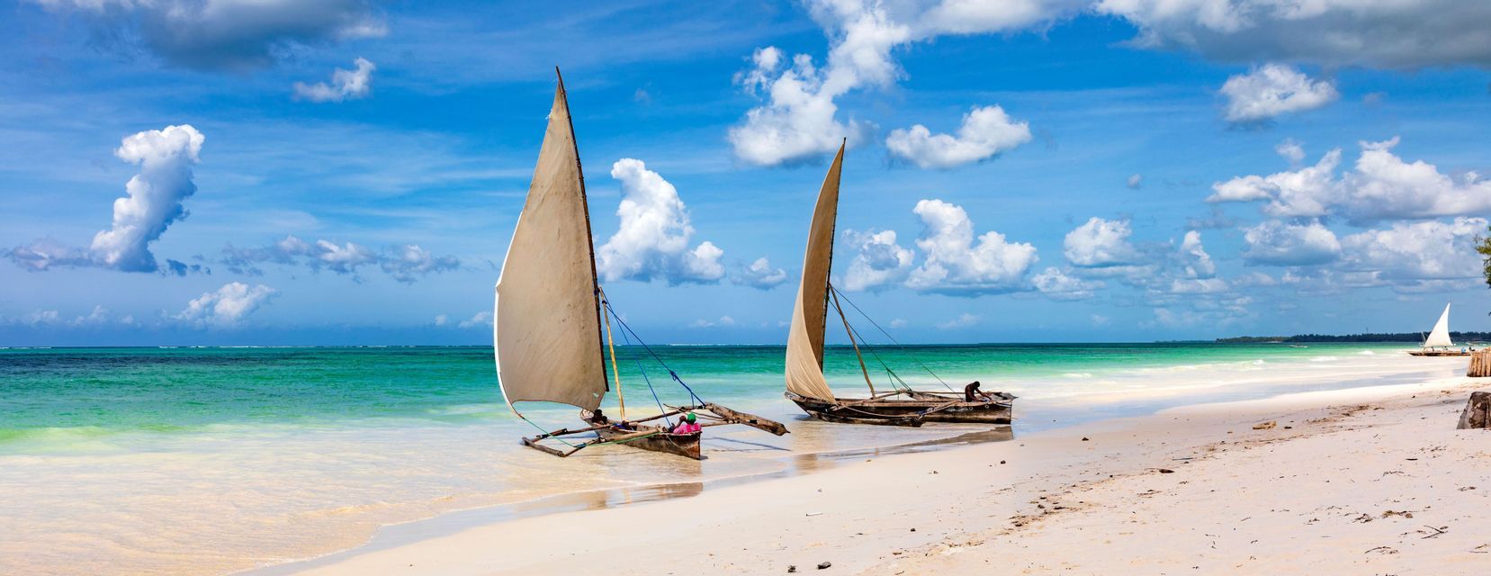 Dos veleros tradicionales con velas de color beige descansan en una playa de arena blanca junto a aguas turquesas claras bajo un cielo azul con nubes blancas.