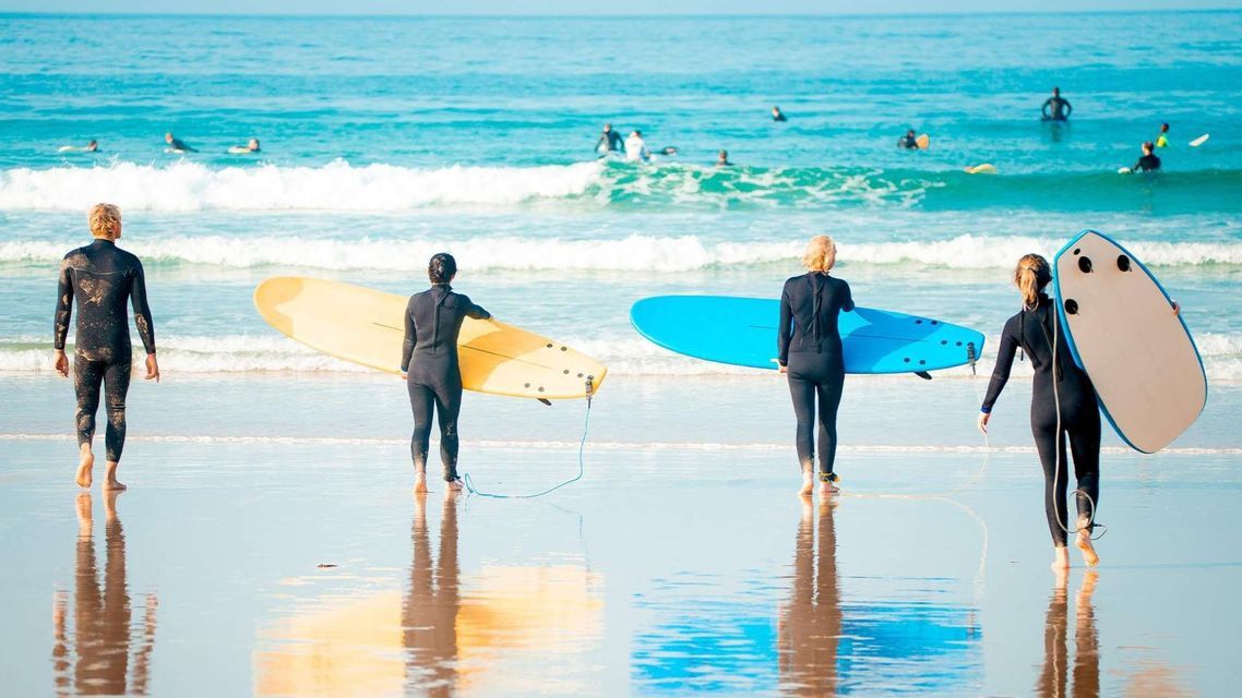 Two surfers in wetsuits from a WeRoad group trip stand on a wet beach, facing the ocean with a yellow surfboard.