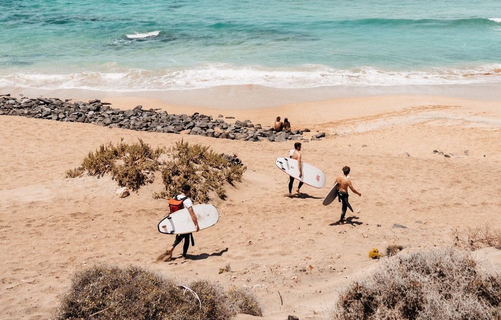 A WeRoad group trip of three surfers carrying their boards as they walk across a sandy beach towards the turquoise ocean.