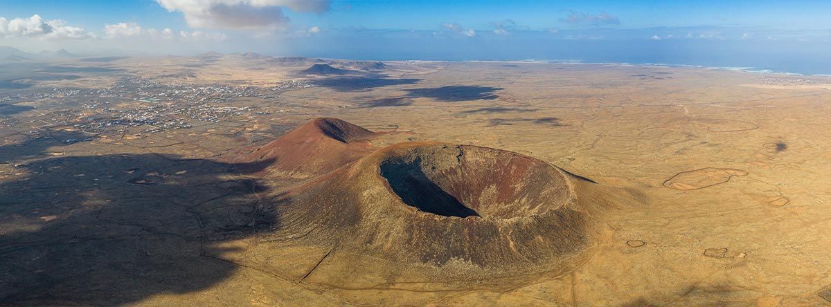 An aerial view of a large volcanic crater in a vast, arid landscape with a small town and the ocean in the distance.