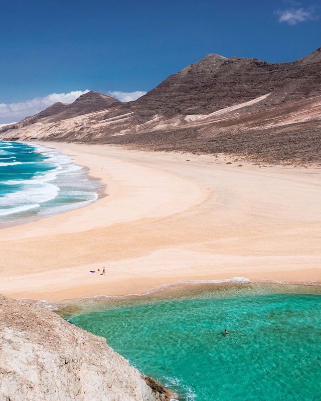 A high-angle view of a vast sandy beach with turquoise water curving at the base of arid mountains under a blue sky.