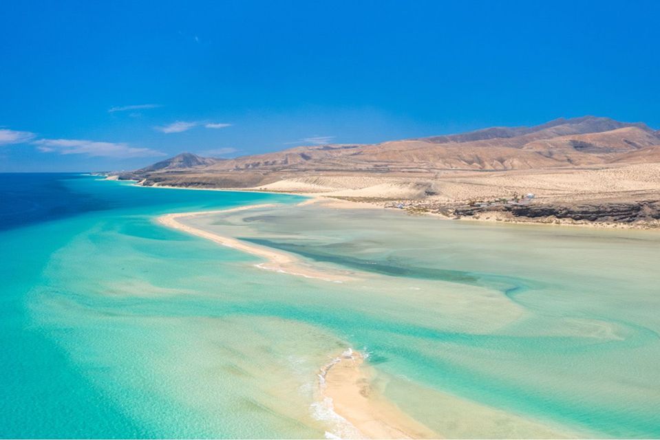 Aerial view of a sandbar extending into clear turquoise water next to a dry, hilly coast under a deep blue sky.