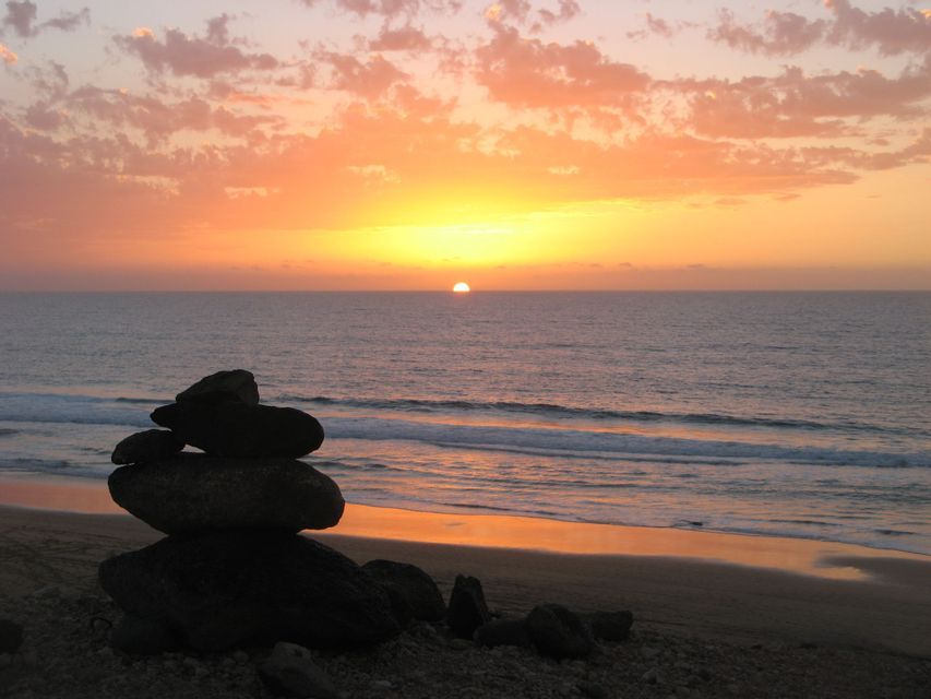 A cairn of stacked rocks sits on a beach, silhouetted against the setting sun over the ocean.