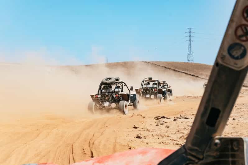 Dune buggies from a WeRoad group trip drive in a convoy across a sandy desert, kicking up dust under a bright blue sky.