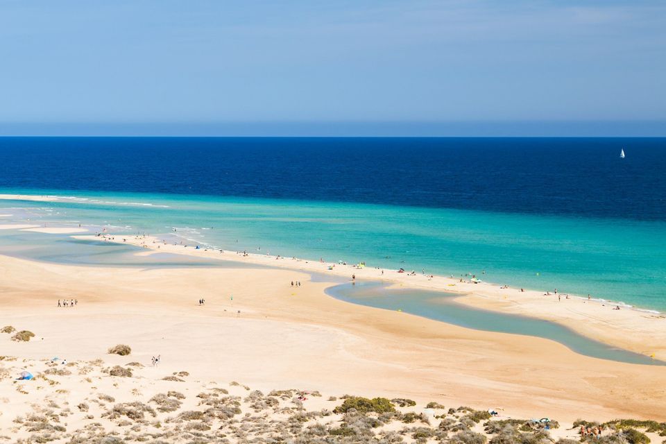 Una vasta spiaggia sabbiosa con persone lungo la riva dove l'acqua turchese incontra l'oceano blu profondo.