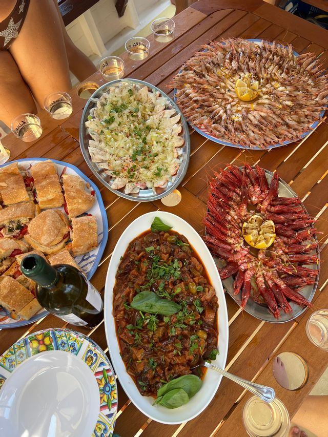 Un pasto all'aperto su un tavolo di legno con vassoi di gamberetti, panini, stufato di verdure e una bottiglia di vino