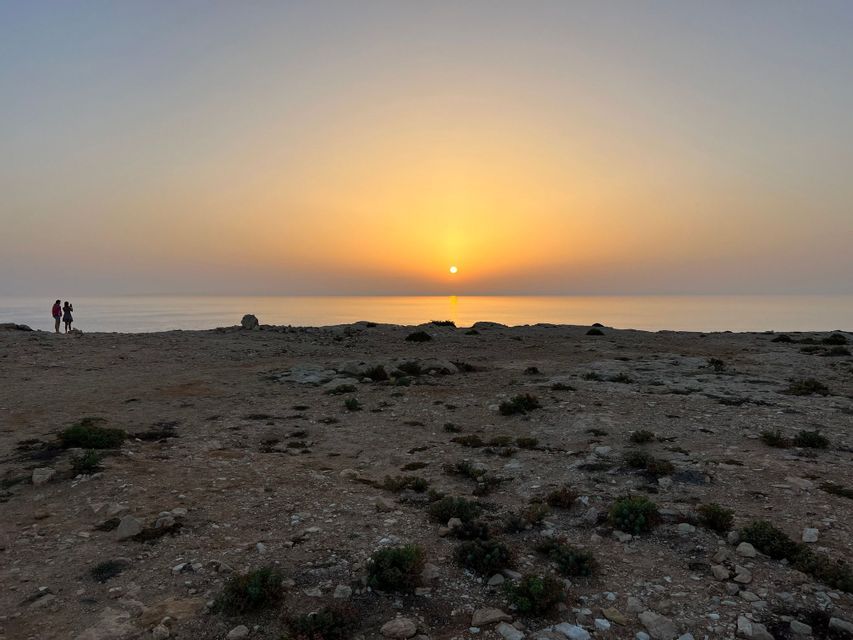 Due persone si trovano su una scogliera rocciosa, ammirando il tramonto sull'oceano calmo.