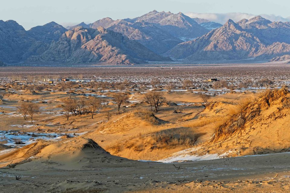 Des dunes de sable doré parsemées de neige recouvrent le fond d'une vallée désertique, avec une chaîne de montagnes accidentées en toile de fond au lever du soleil.