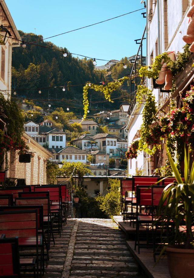 A cobblestone alley lined with red chairs from an outdoor cafe ascends a hill with stone houses and string lights.